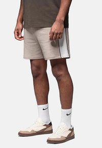 Shorts de sport beige avec une bande noire sur le côté, portés avec des chaussettes blanches et des baskets beige-brun. Coupe décontractée avec une texture douce.