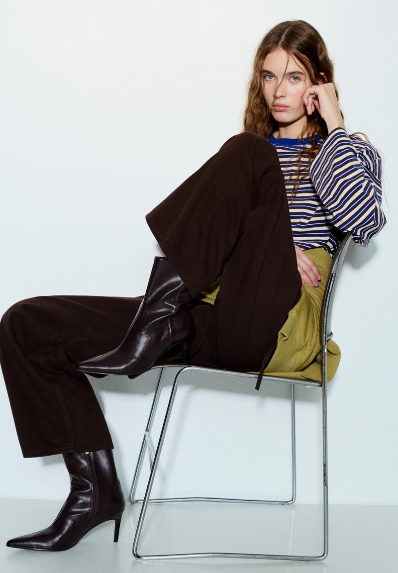 Femme aux longs cheveux ondulés assise de profil sur une chaise en métal, portant une chemise rayée à manches longues, un pantalon large foncé et des bottes pointues en cuir foncé.
