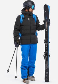 Skiausrüstung mit einer schwarzen Daunenjacke, blauen Skihosen, einem schwarzen Helm und Sonnenbrille. Hält schwarze Skier mit blauen Akzenten und Trekkingstöcke.