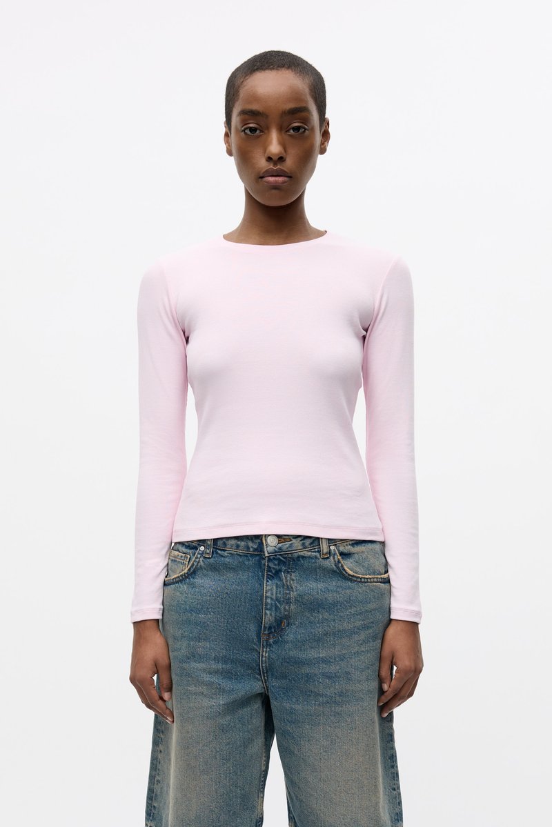 Camicetta a maniche lunghe di un delicato rosa chiaro con scollatura rotonda, realizzata in un tessuto morbido e liscio, abbinata a jeans in denim blu dal taglio morbido.