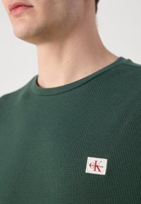 Donkergroen geribbeld sweatshirt met een ronde hals. Bevat een vierkant label met "calvinKlein" in het rood op een witte achtergrond.