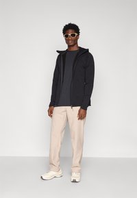 Crna zip-up hoodie s mekanom teksturom, nosi se preko tamno sive majice. Uparen s svijetlosmešavim hlačama i bijelim tenisicama. Okviraste naočale.