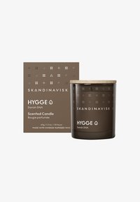 Bougie parfumée dans un pot en verre marron foncé avec un couvercle en bois. L'emballage présente des motifs géométriques et du texte blanc sur un fond marron.