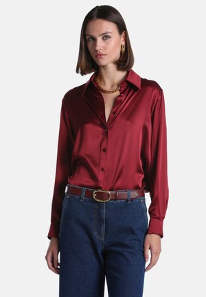 Femme portant une chemise bordeaux en satin à boutons, un jean bleu, une ceinture assortie, un collier chaîne en or et des boucles d’oreilles créoles en or sur fond blanc.