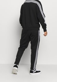 Czarny dres z kurtką na zamek z trzema białymi paskami na ramionach i pasującymi spodniami z detalami w paski, połączony z czarnymi butami.