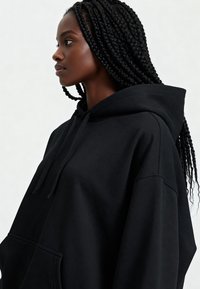 Čierna nadrozmerná mikina s kapucňou a vreckom typu klokanka, vybavená šnúrkami a textúrovaným povrchom. Bočný profil modelu so zapletenými vlasmi.