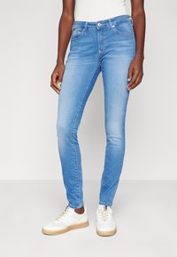 Vaqueros pitillo de mezclilla azul claro con una textura suave, diseño de cinco bolsillos, cierre de botón y un sutil efecto de desgaste. Combinados con zapatillas blancas.