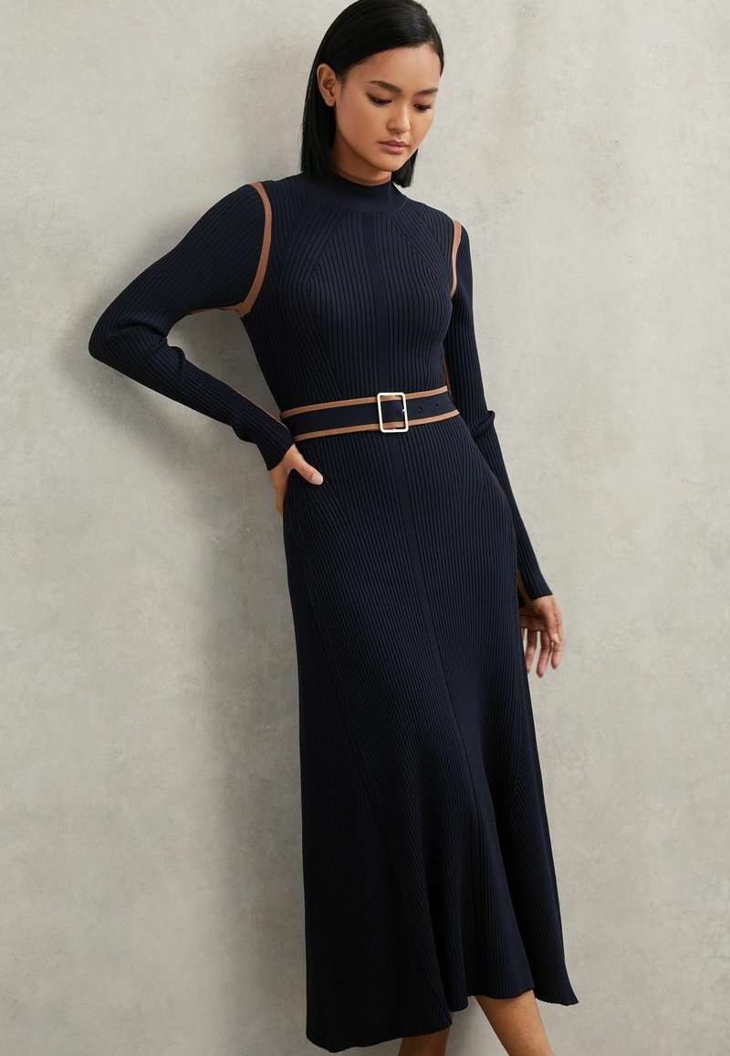 Marineblauwe geribbelde jurk van gebreide stof met lange mouwen en een taille met ceintuur. Voorzien van contrasterende bruine schouderaccenten en een uitlopende zoom.