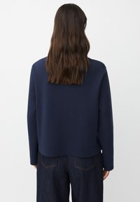 Osoba z długimi brązowymi włosami, ubrana w granatową bluzę z długim rękawem i ciemne dżinsy, zwrócona plecami w stronę jednolitego tła.