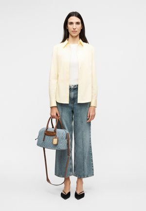 Kvinne med langt mørkt hår iført en lys gul skjorte med knapper, vide jeans, svarte sko, holder en blå mønstret håndveske med brune stropper.