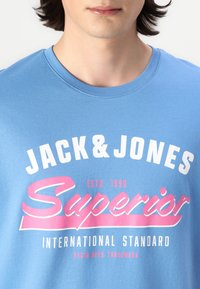 Šviesiai mėlynas medvilninis marškinėlių su trumpomis rankovėmis ir apvaliu iškirpimu. Puošia baltas ir rožinis grafikos tekstas, įskaitant „Jack & Jones“ ir „Superior.“