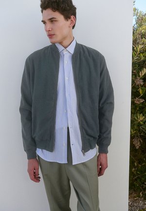 Chaqueta bomber gris con cierre delantero, llevada sobre una camisa de botones a rayas azul claro y blanca y pantalones verde oliva. Textura de tela suave.