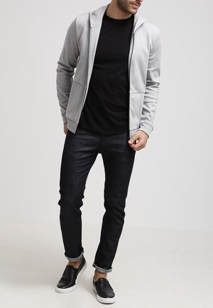 Sudadera con cremallera de color gris claro sobre una camiseta negra, combinada con jeans oscuros y zapatos negros sin cordones. Tejido suave, ajuste casual, diseño minimalista.
