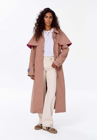 Kobieta z kręconymi włosami, ubrana w długi beżowy płaszcz, białą koszulę, kremowe spodnie i buty w cętki, stojąca na tle białej ściany.