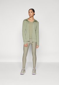 Conjunto desportivo verde-oliva composto por um hoodie ajustado de mangas longas, top correspondente e leggings, complementado com ténis cinza claro.