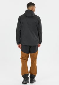 Mand stående med ryggen mod, iført en sort hættetrøje og brune og sorte bukser med sorte vandresko på en hvid baggrund.