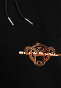 Zwarte hoodie met een glanzend oranje logo van een gestileerd aapje, met reflecterende tekst "BLCKBNNS" eronder. Koordjes met zilveren uiteinden.