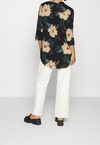 Blusa nera a fiori con fiori beige e foglie verdi, maniche a tre quarti, orlo curvo, abbinata a pantaloni bianchi a gamba larga e scarpe nere con zeppa.