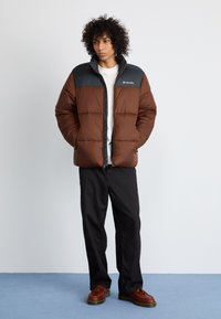 Chaqueta de abrigo marrón con un panel superior negro, que presenta un cierre frontal, un ligero brillo y costuras horizontales. Combinada con pantalones negros y zapatos marrones.