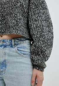 Grauer und schwarzer Strickpullover mit einem verkürzten Design, das ein strukturiertes Muster und übergroße Ärmel aufweist, kombiniert mit hellblauen, hochtaillierten Jeans.