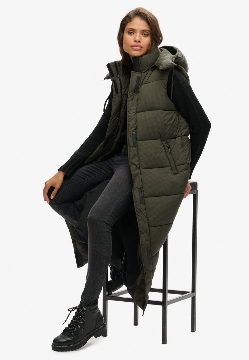 Langer, olivgrüner Daunenweste mit Kapuze, die mit einem Reißverschluss versehen ist und gepolsterte Abschnitte aufweist. Kombiniert mit schwarzen Jeans und Schnürstiefeln.