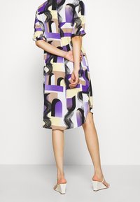 Femme portant une robe longueur genou avec des motifs abstraits en violet, beige et noir, se tenant les mains derrière le dos, sandales compensées blanches.