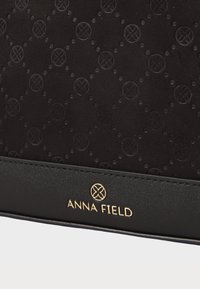 Čierna textúrovaná tkanina s embosovaným kruhovým vzorom a zlatým logom "ANNA FIELD" na hladkom čiernom koženom prúžku.