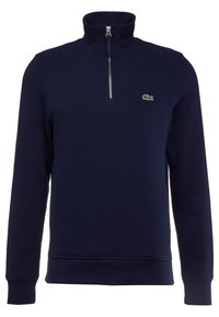 Marinblå zip-tröja med hög krage, räfflad textur och en liten grön Lacoste-logotyp på bröstet. Klassisk passform.