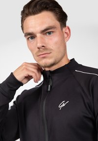 Zwarte sportieve zip-up jas met een hoge kraag, voorzien van witte accenten en een logo op de borst; de stof lijkt glad en aansluitend.