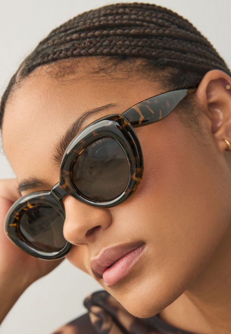 Tortoiseshell sunglasses with oversized, rounded frames, dark lenses, and subtle gold accents on the temples. Smooth plastic texture.