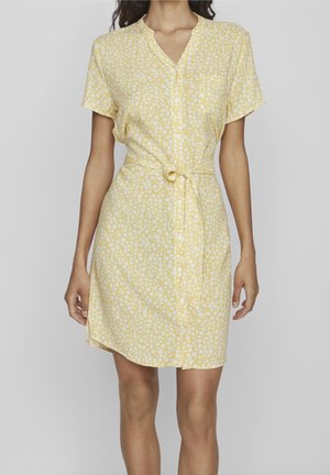 Kvinde iført gul kjole med korte ærmer, knælængde, hvide abstrakte pletter, knaplukning foran, krave og bælte i taljen, stående mod en ensfarvet baggrund.