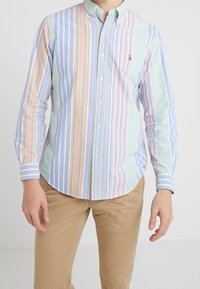 Camicia con colletto e bottoni in righe verticali pastello di blu, verde, rosa e arancione, con un piccolo logo sul petto e maniche lunghe.