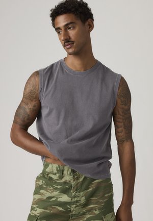 Άνδρας με τατουάζ φορώντας αμάνικο γκρι πουκάμισο και καμουφλάζ cargo σορτς, κοιτάζει προς τα πλάγια με το ένα χέρι στην τσέπη.