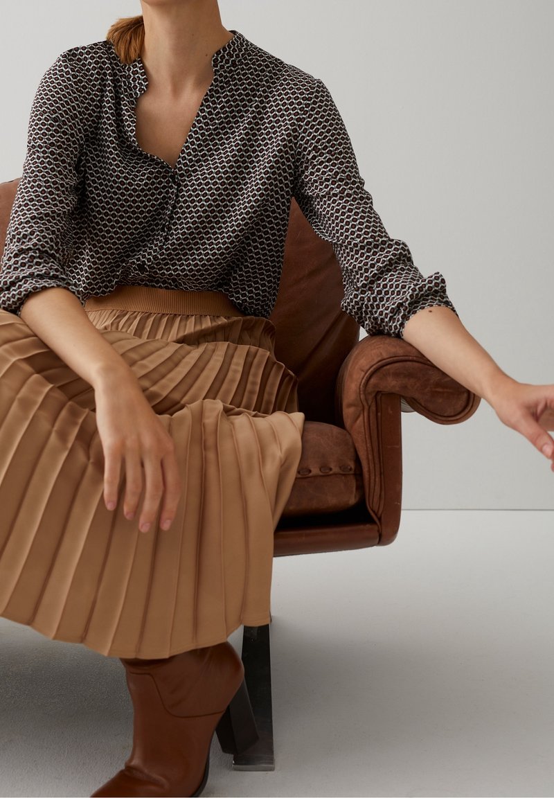 Blusa a righe in bianco e nero con colletto alla cinese, abbinata a una gonna plissettata beige e stivaletti con tacco marroni, seduta su una sedia in pelle marrone.