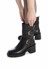 Botas de tobillo de cuero negro con dos hebillas doradas, tacones gruesos y una suela texturizada. Modelo ajustando la parte superior con medias de red visibles.
