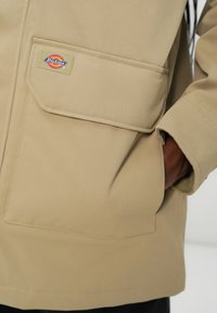 Bežna jakna iz teksturiranega materiala z veliko sprednjo žepom, s pisanem logotipom Dickies, prišitim na pokrovu žepe.