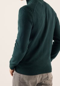 Donkergroen gebreide trui met geribde kraag, gestructureerde schouderaccenten en nauwsluitende mouwen, gecombineerd met lichtgrijze broek.