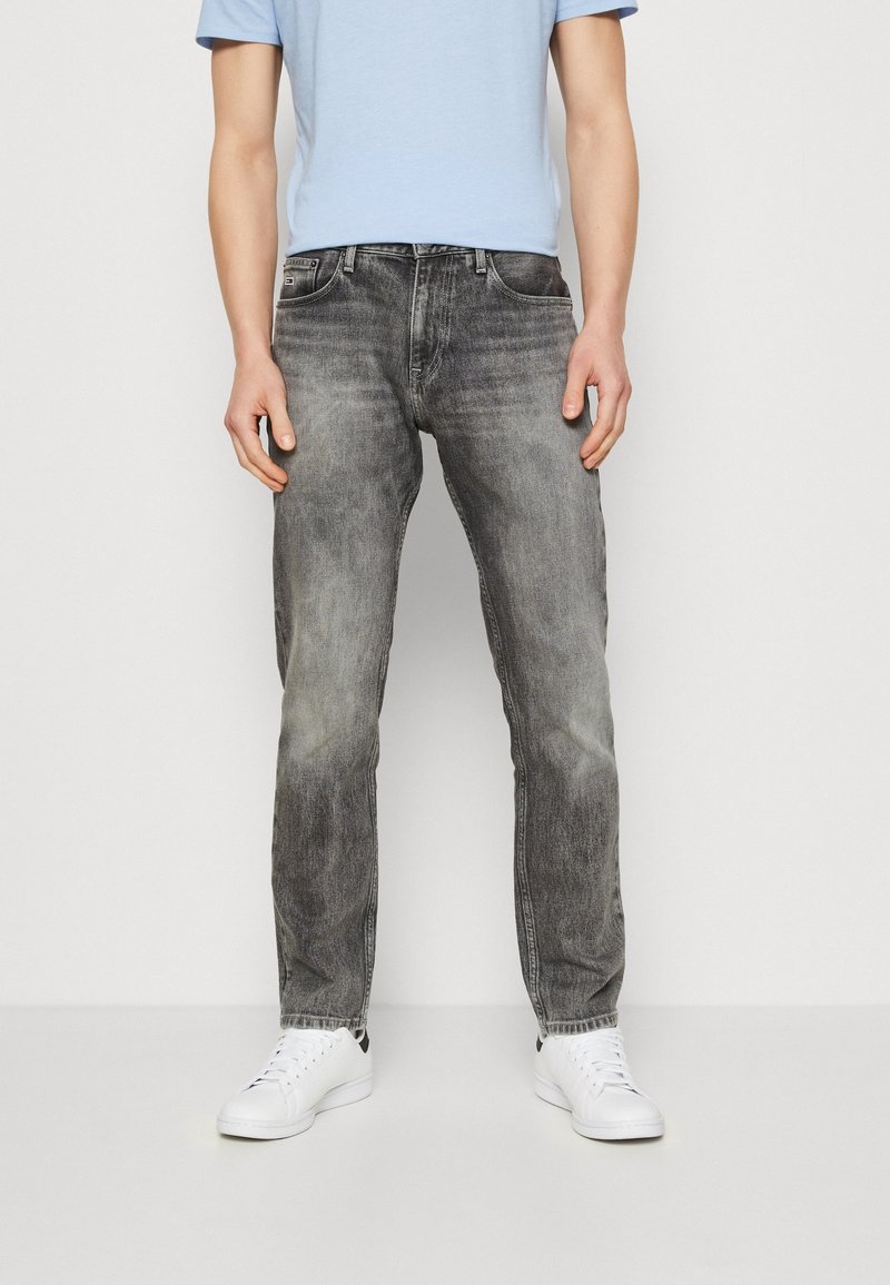 Jeans de mezclilla gris con acabado deslavado, diseño clásico de cinco bolsillos y corte recto, combinados con zapatillas blancas y una camisa azul claro.