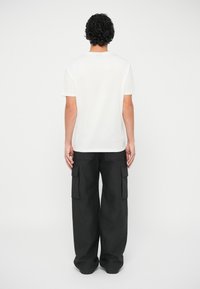 Homem com cabelo encaracolado vestindo uma camiseta branca simples e calças de cargo pretas largas, de pé de costas num fundo branco.