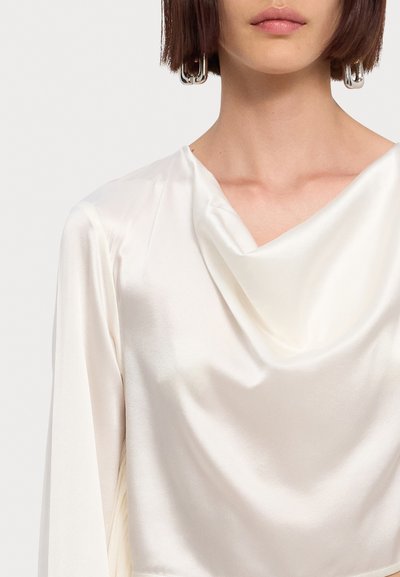 Άσπρη σατέν μπλούζα με ντραπέ λαιμόκοψη και μακριά μανίκια. Το ύφασμα είναι λείο και γυαλιστερό, ενισχύοντας τη κομψή σιλουέτα. Ορατά ασημένια σκουλαρίκια σε σχήμα κρίκου.