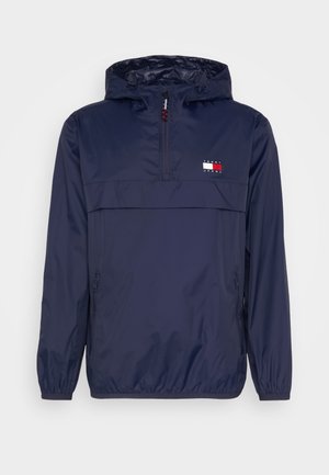Veste coupe-vent à capuche bleu marine en tissu léger et résistant à l'eau, dotée d'une fermeture éclair à mi-hauteur et d'un logo sur la poitrine.