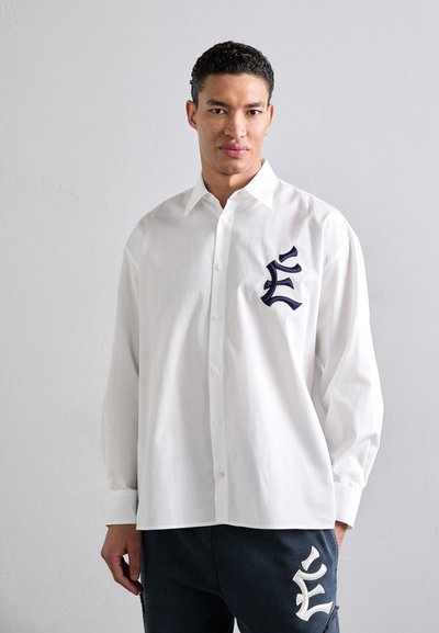 Camicia bianca con bottoni realizzata in cotone, caratterizzata da una grande lettera blu ricamata sul petto, con una vestibilità comoda e maniche lunghe.