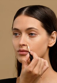 Concealer wand applying light beige product under the eye. Smooth skin texture with neutral background. Hand holding applicator with black casing.