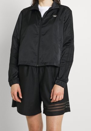 Person wearing black zip-up jacket with Adidas logo and black shorts featuring three sheer stripes on left leg.