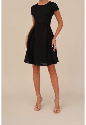 Frau trägt ein knielanges schwarzes strukturiertes Kleid mit Kappärmeln und beige High-Heel-Sandalen, steht vor einem schlichten Hintergrund.