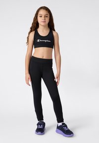 Fille jeune debout, portant un soutien-gorge de sport Champion noir, un legging noir, des chaussettes blanches et des baskets noires avec des semelles et des lacets violets.