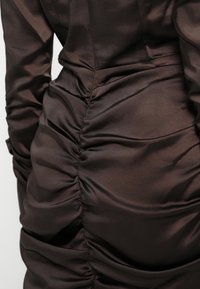 Rochie din satin maro cu un design plisat pe spate, cu textură netedă și luciu subtil. Croiala adaptată evidențiază forma.