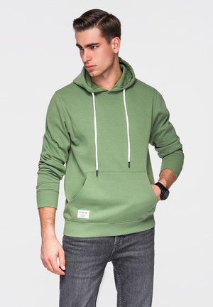 Jeune homme portant un sweat à capuche vert avec des cordons blancs et un jean gris, main gauche dans la poche, regardant vers sa gauche sur un fond clair.