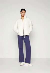 Donna in piedi con le mani in tasca, che indossa un giubbotto imbottito bianco, pantaloni larghi viola e sneakers bianche, su uno sfondo neutro.