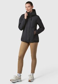 Črna lahka jakna z kapuco, z zadrgo zaprtimi žepi, elastičnimi manšetami in prilegajočo silhueto. Skombinirana je s bež hlačami in copati.